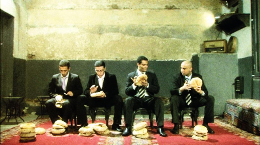 EKSEMPEL Adel Abidin, Bread of Life, 2009. Still: Adel Abidin I videoværket Bread of Life af Adel Abidin sidder fire musikere iført sorte jakkesæt på række og spiller på tørre brød.